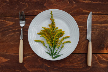 Yellow field flower with green leaves in a white plate with a knife and fork on a brown wooden table top view, diet concept, healthy food, practical dinner