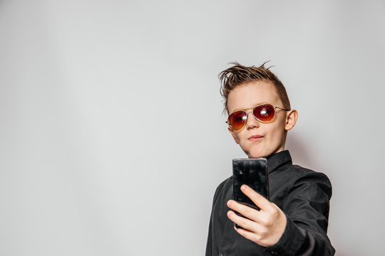 a fashionable guy in sunglasses and a black shirt with a cool hairstyle looks at the phone and photographs himself on a white background.