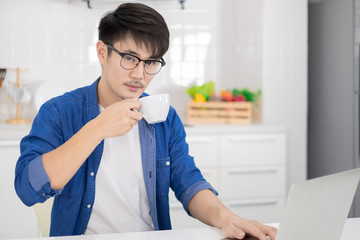 Asian freelance young man working from home with coffee using laptop in white kitchen