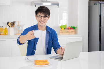 Fototapeta na wymiar Asian freelance young man working from home with breakfast bread and coffee using laptop in white kitchen