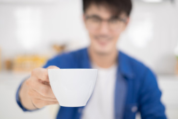 Asian man holding a cup of coffee and sitting in white kitchen. Selective focus on mug