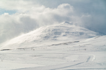 A snow covered mountain during incredible winds at its peak. Snow is blowing from its top, merging with the clouds around it, yet the colors are warm.
