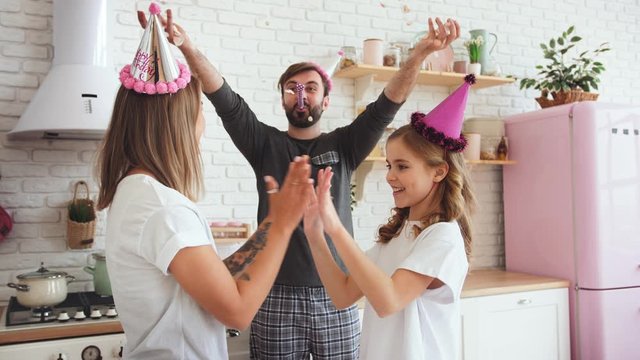 Happy family in pajamas with a daughter dancing and celebrating birthday in kitchen during online birthday party. Dad is throwing confetti, slow motion