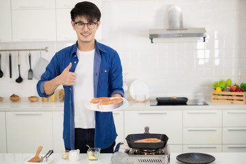 Asian man coking salmon steak with grill pan in white kitchen.