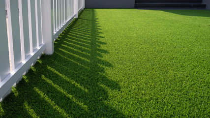 Sunlight and shadow of white wooden fence on green artificial turf surface in front yard of home,...