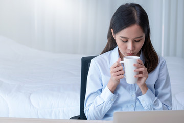 Front view of pretty business girl with blue shirt drink coffee during work at home in bedroom with morning light.
