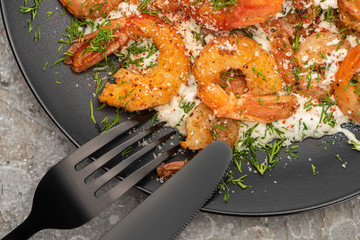close up view of fried shrimps in sauce with dill in black plate on grey concrete background with cutlery