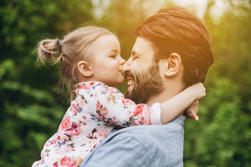 stylish bearded father with his little daughter in his arms against the backdrop of green trees on a walk in the park hugging and kissing a baby - obrazy, fototapety, plakaty