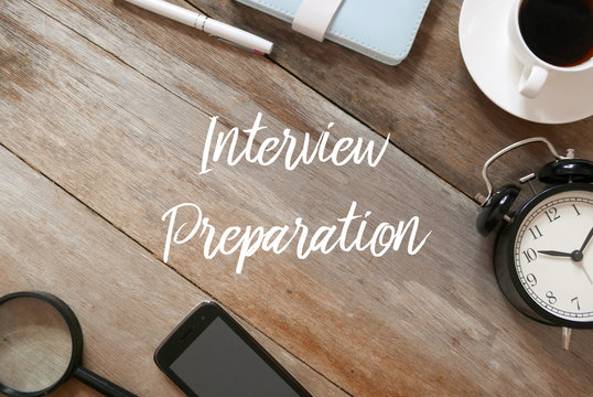 Top view of magnifying glass,mobile phone,vintage table clock, a cup of coffee,notebook and pen on wooden background written with Interview Preparation.