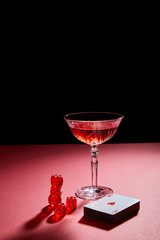 glass of cocktail near deck of cards and dice on red surface isolated on black