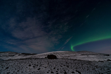 Fototapeta na wymiar Northern lights in Iceland on dark blue sky with a hint of clouds over a snow covered barren mountain landscape with a large rock in the foreground.