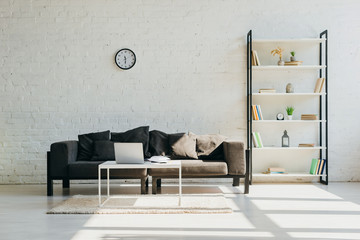 living room with grey sofa, clock, shelf and table with laptop in sunlight