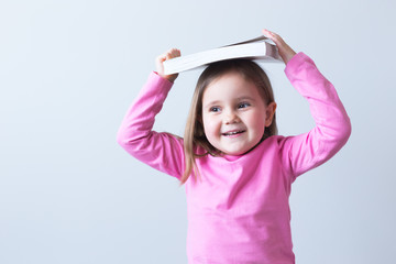 Portrait of a white girl 4 years old. Pink clothes looking to the side. Holds a book on her head with her hands. Gray background. Selective focus.