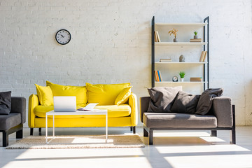 living room with yellow sofa, grey armchairs, shelf, clock and laptop in sunlight