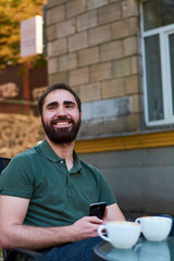 Portrait of young guy with dark beard, sitting in coffee shop on summer terass and drinking coffee in green shirt.