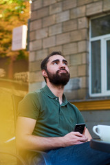 Portrait of young guy with dark beard, sitting in coffee shop on summer terass and drinking coffee in green shirt.