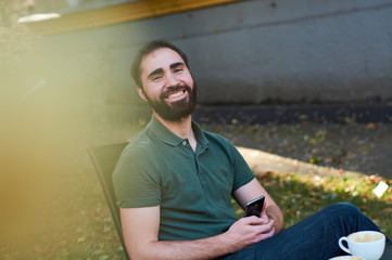 Portrait of young guy with dark beard, sitting in coffee shop on summer terass and drinking coffee in green shirt.