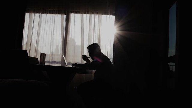 Silhouette of a man who is drinking coffee while working using a laptop in a modern room or at home with a window background that is illuminated by slightly blinding natural glare. Work from home.