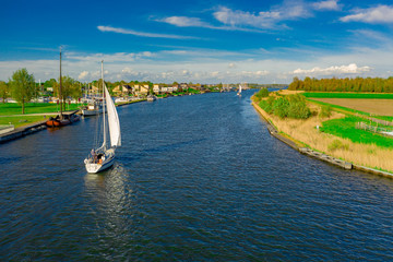 Aerial drone photo of sailboats passing through a canal in the Netherlands with beautiful modern houses in the background with a beautiful blue sky and a few clouds