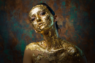 Girl with a mask on her face made of gold leaf. Gloomy studio portrait of a brunette on an abstract...