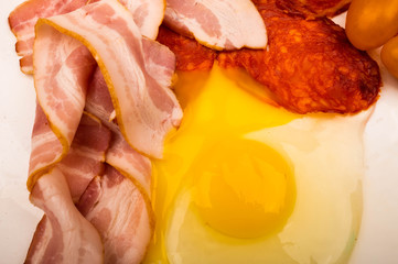A broken chicken egg without a shell, slices of sausage and bacon and tomatoes on a white background. Close up.