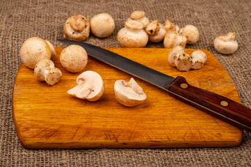 Halves of cut young mushrooms and a knife with a wooden handle on a wooden cutting Board and mushrooms scattered on a background of coarse homespun fabric. Close up.