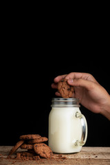 A few stacks of cookies, a glass of fresh milk and a hand holding a cookie that wants to mix it with milk cream isolated on black background
