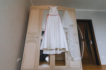Fototapeta na wymiar Wedding white long dress of the bride hangs on a hanger, on a wooden wardrobe with a mirror in the hotel room. Photography, concept.