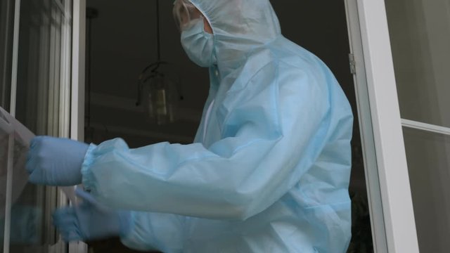 COVID-19 disinfection. Worker in a protective blue overalls removes DISINFECTION sign after disinfection in the room. Disinfector in a protective suit and mask sprays disinfectants in house or office.