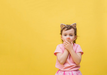 The frightened girl covers her mouth with her hands on a yellow background. A little girl with a cat-ear headband is surprised. Cute girl in a pink t-shirt with a pocket.