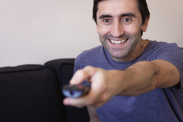 Smiling man pointing with tv remote control in living room. Happy person doing channel surfing on...