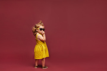 Cool stylish little girl in yellow dress and sunglasses and sunglasses isolated at the pink background. Child fashion. Copy space