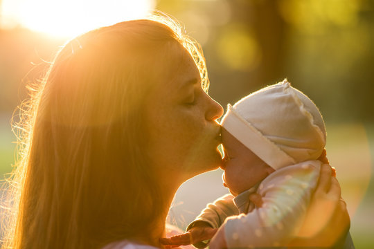 A young mother kisses her newborn baby in a summer sunset park. Maternity Happiness