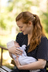 A young mother with a small newborn in her arms sits on a bench in a summer park. Happiness...