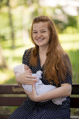 A young mother with a small newborn in her arms sits on a bench in a summer park. Happiness...