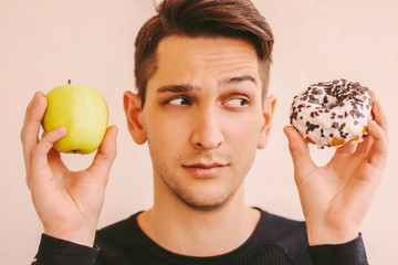 Portrait of uncertain young sports man holding sweet donut and fresh apple in hands. Funny fitness man deciding what to choose: sweet sugar donut or healthy fruit. Sports nutrition motivation, dieting