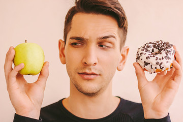 Portrait of funny uncertain fitness man hold fresh apple and sweet donut snack in hands. Young...