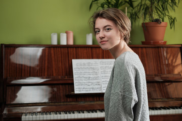 Young woman smiling sitting near acoustic piano with music sheet. Home islolation.