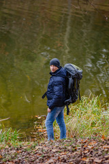 Tourist in a jacket and jeans looks at the camera near the river. Autumn landscape background.