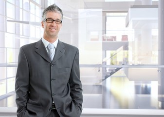 Mature caucasian financial business expert businessman standing at high-tech office. Smiling, looking at camera, suit and tie, hands in pocket, wearing glasses, copyspace.