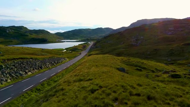 Aerial drone flight past a young girl standing on a hill in the middle of a beautiful valley. Road passes stones and green hills in area of lake Ivlungsvotni in Norway. Taken in the summer at sunset