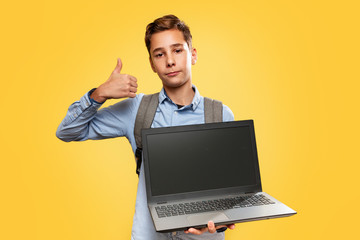 Concept of modern technologies and education. Caucasian teen boy holding open laptop and showing thumbs up. Yellow background. Copy space