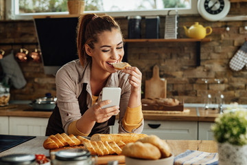 Happy woman eating slice of bread while using smart phone in the kitchen.