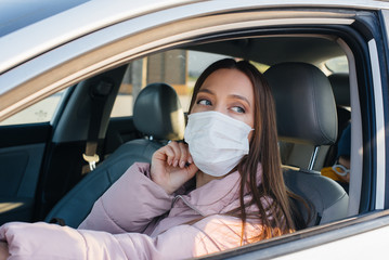Fototapeta na wymiar A young girl sits behind the wheel in the car in the mask during the global pandemic and coronavirus. Quarantine