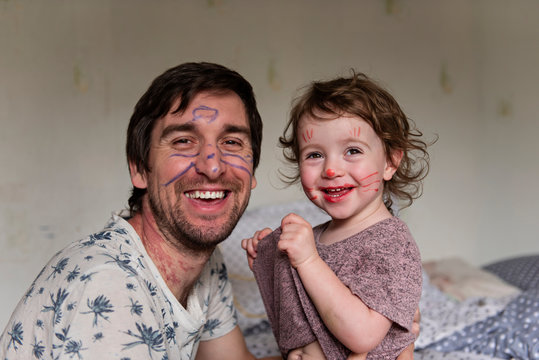 A young man, a father, and a beautiful little girl, a daughter, drawing markers on each other’s faces, laughing and looking at the camera