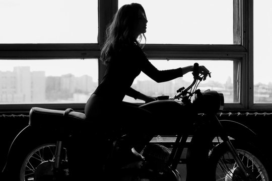 A silhouette of a woman with long hair sitting on a motorcycle. Black and white photo