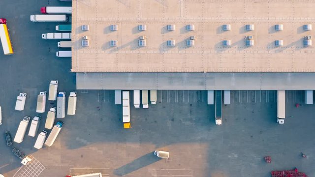 Aerial hyper lapse (hyperlapse - time lapse) of the large logistics park with warehouse, loading hub and semi trucks with cargo trailers standing at the ramps for load/unload goods. Top down view