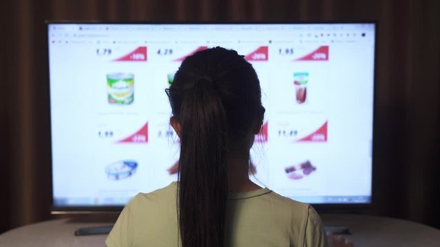 Back view of female sitting at big screen PC and chooses food and other products for an online home order.