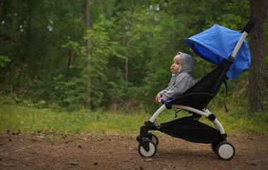 Small thoughtful child in sweatshirt hood on head sitting in baby stroller in green park, little travelling child