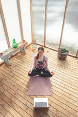 Young woman is seated in lotus pose at her home yoga studio. She is focused on listening to the guided meditation. Care about our mental health in quarantine. Cosy, bright and wooden interior. 
